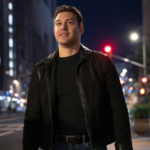 Man wearing a black leather jacket standing on a city street at night with blurred lights in the background