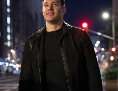 Man wearing a black leather jacket standing on a city street at night with blurred lights in the background