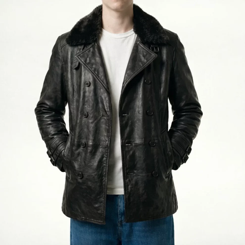A man wearing a black double-breasted leather jacket with a fur collar, styled with a white shirt and blue jeans, standing against a plain background.
